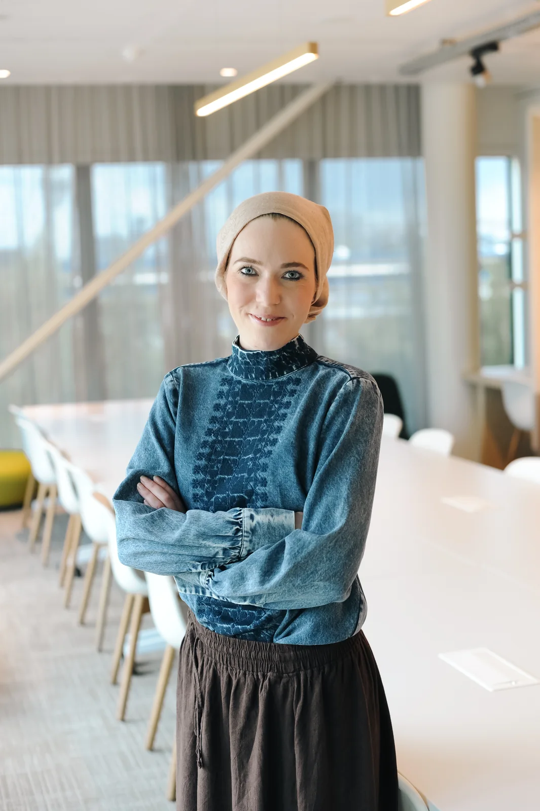 Portrait of Daphne standing with arms folded beside a long meeting-room table, looking directly at the camera.